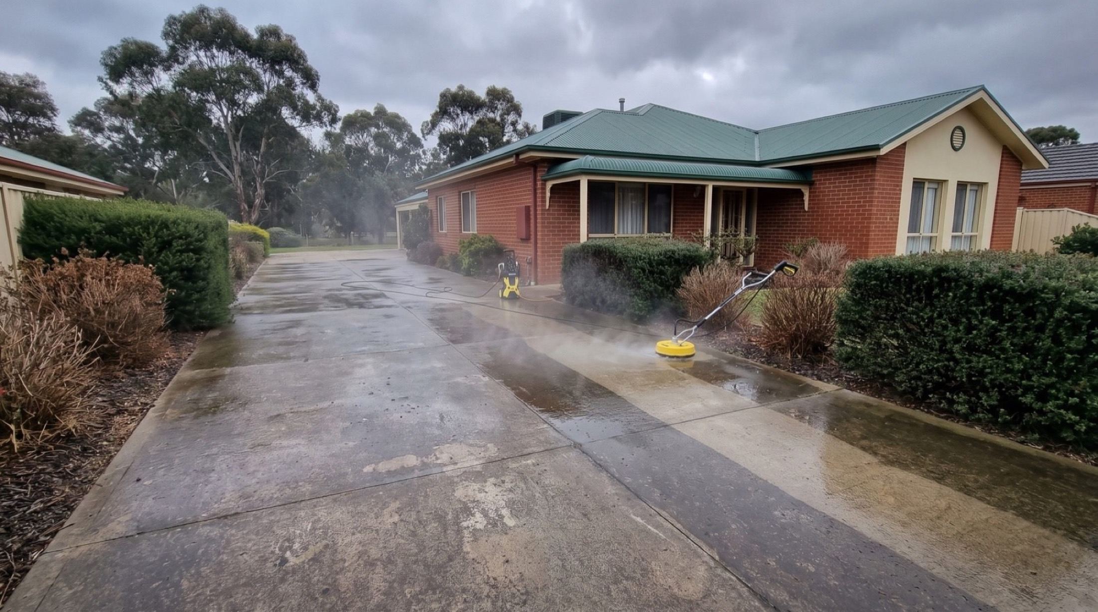 A concrete driveway being pressure washed beside a brick home and clipped hedges