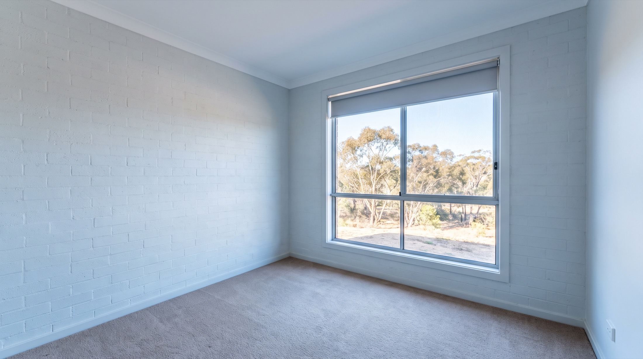 An empty bedroom with bare carpet, plain walls and a window with the blind half raised